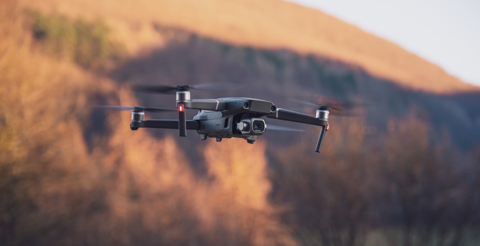 A quadcopter drone flying in front of a sunlit hillside at sunset